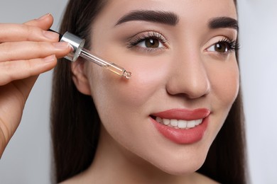 Photo of Happy young woman applying essential oil onto face on light grey background, closeup