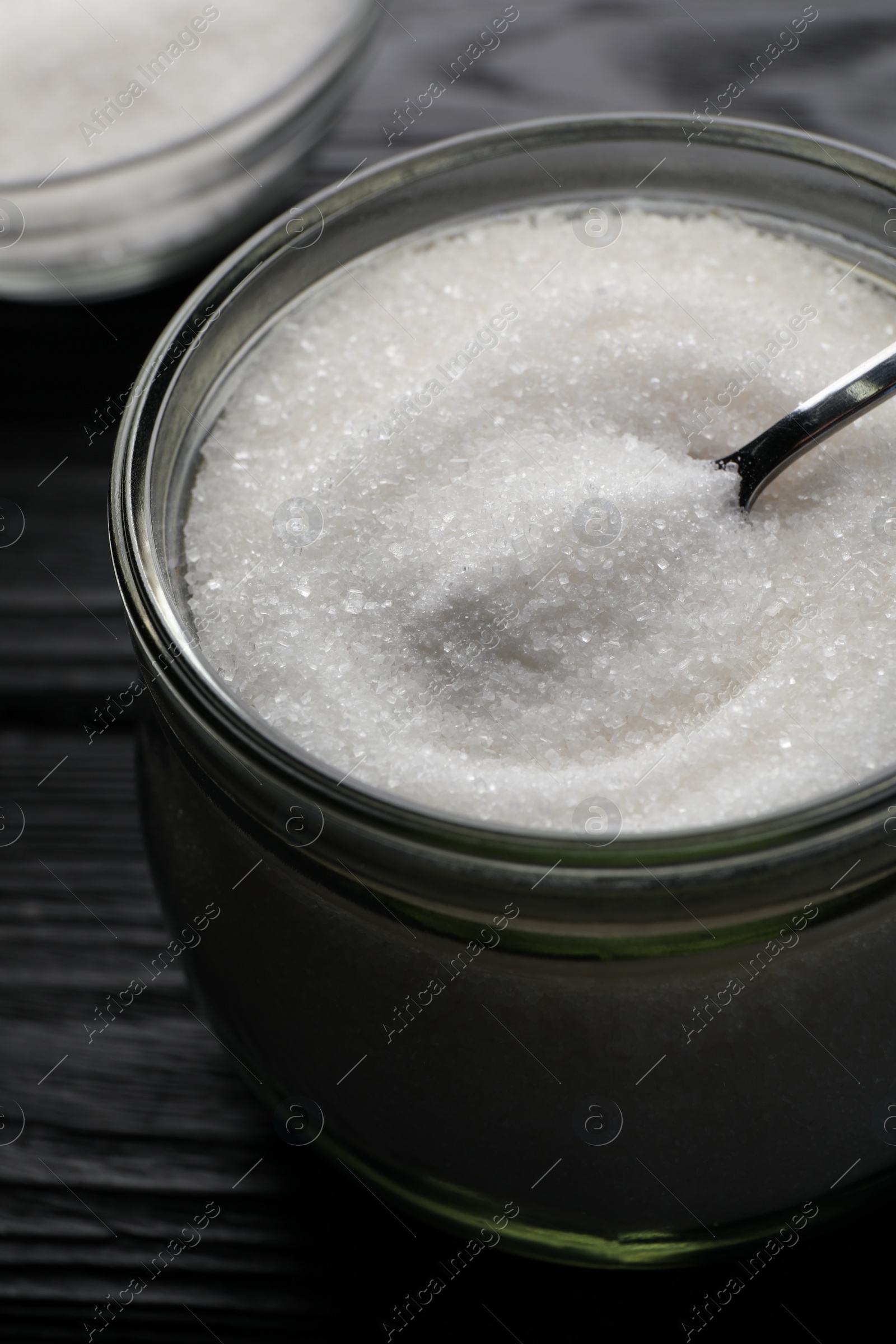 Photo of Granulated sugar and spoon in glass jar on black wooden table, closeup