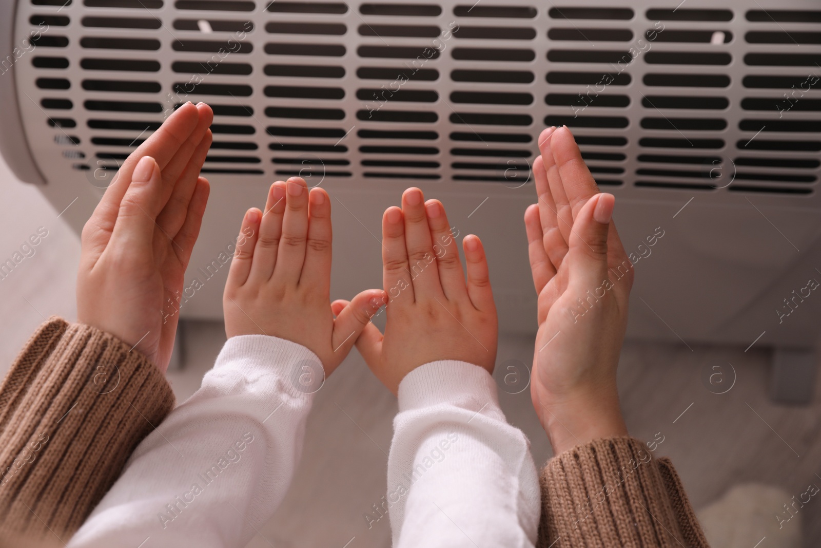 Photo of Mother and child warming hands near electric heater at home, closeup