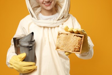 Beekeeper in uniform holding smokepot and hive frame with honeycomb on yellow background, closeup