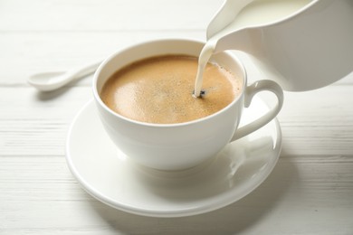 Pouring milk into cup of hot coffee on white wooden table