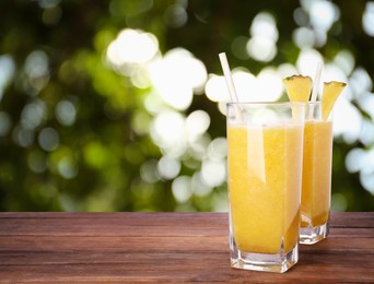 Image of Tasty pineapple smoothie in glasses on wooden table against blurred background, space for text