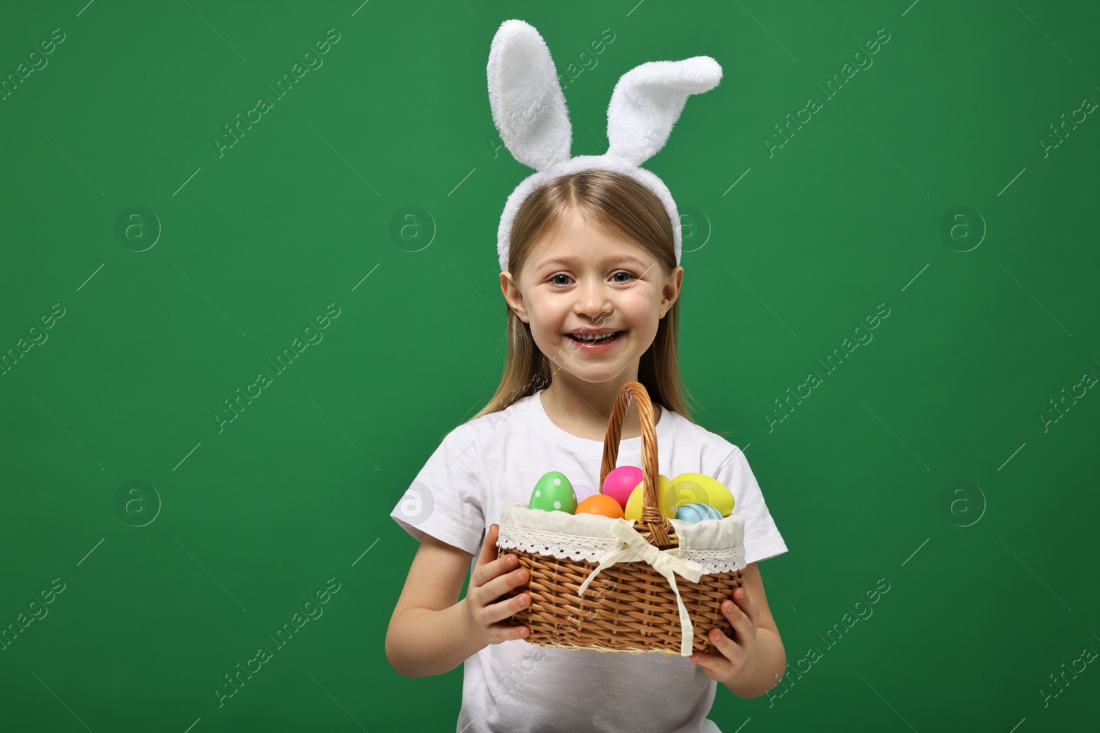 Photo of Easter celebration. Cute girl with bunny ears holding basket of painted eggs on green background