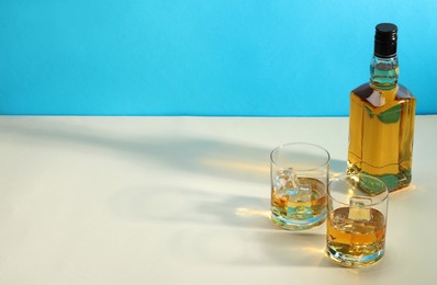 Photo of Whiskey with ice cubes in glasses and bottle on white table against light blue background, space for text