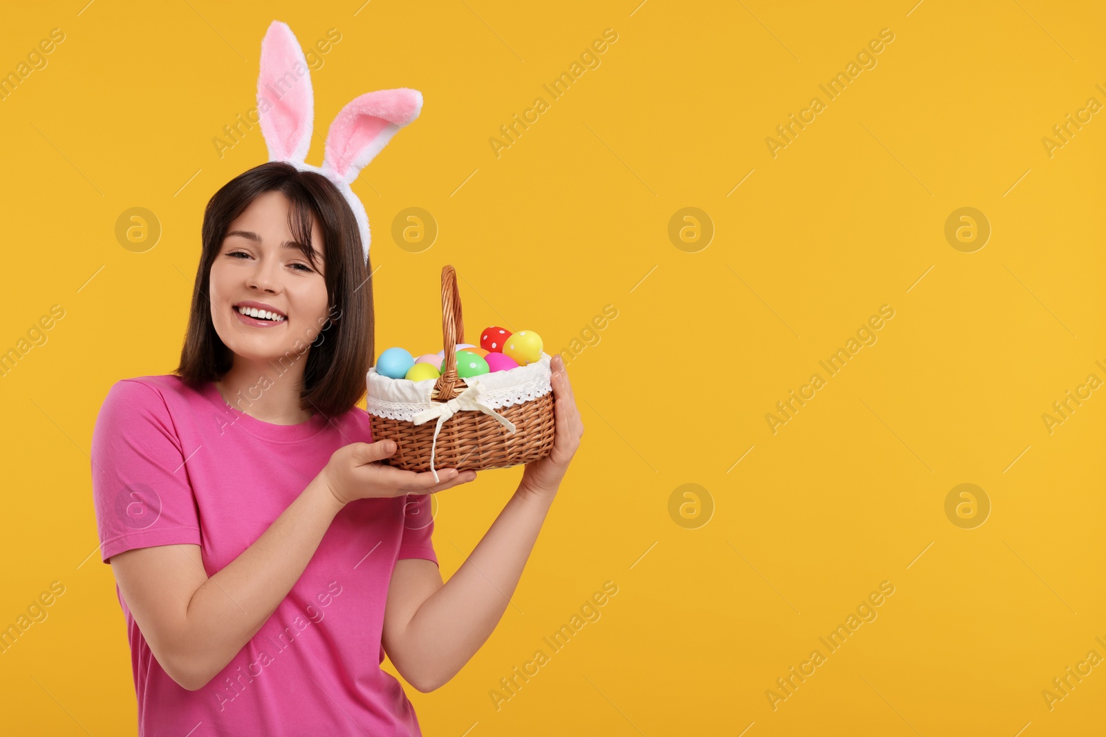 Photo of Easter celebration. Happy woman with bunny ears and wicker basket full of painted eggs on orange background, space for text