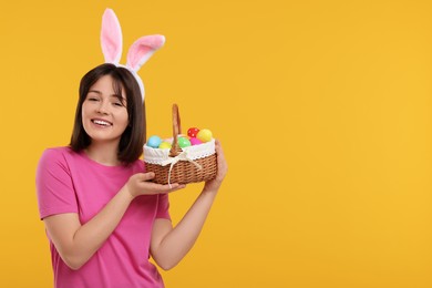 Easter celebration. Happy woman with bunny ears and wicker basket full of painted eggs on orange background, space for text