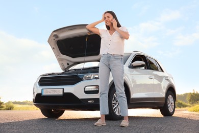 Stressed young woman talking on phone near broken car on roadside, low angle view