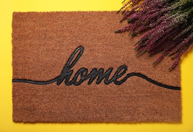 Photo of Doormat with word Home and heather flowers on yellow background, flat lay