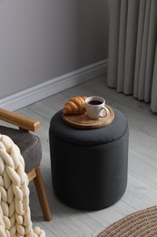 Photo of Tray with cup of tea and croissant on stylish ottoman in room. Interior design
