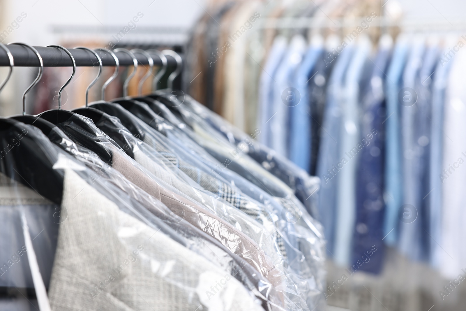 Photo of Dry-cleaning service. Many different clothes in plastic bags hanging on rack indoors, closeup