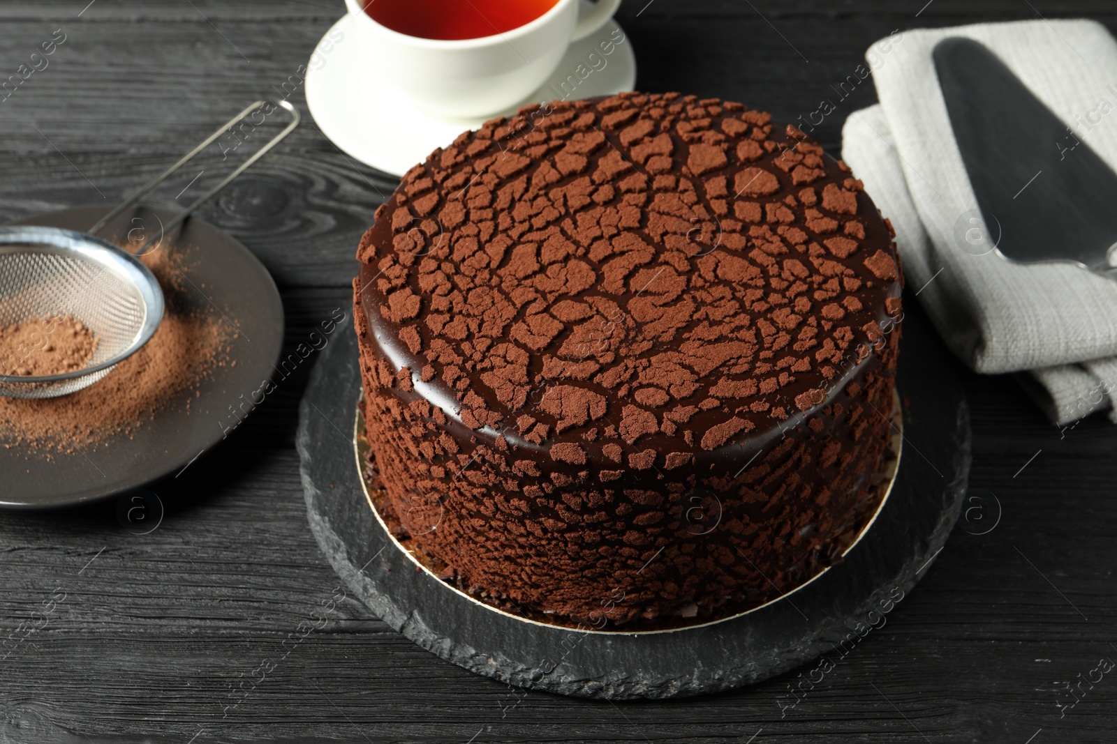 Photo of Delicious chocolate truffle cake, cocoa powder and tea on black wooden table