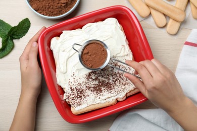 Woman pouring powdered cocoa onto tiramisu cake at wooden table, top view