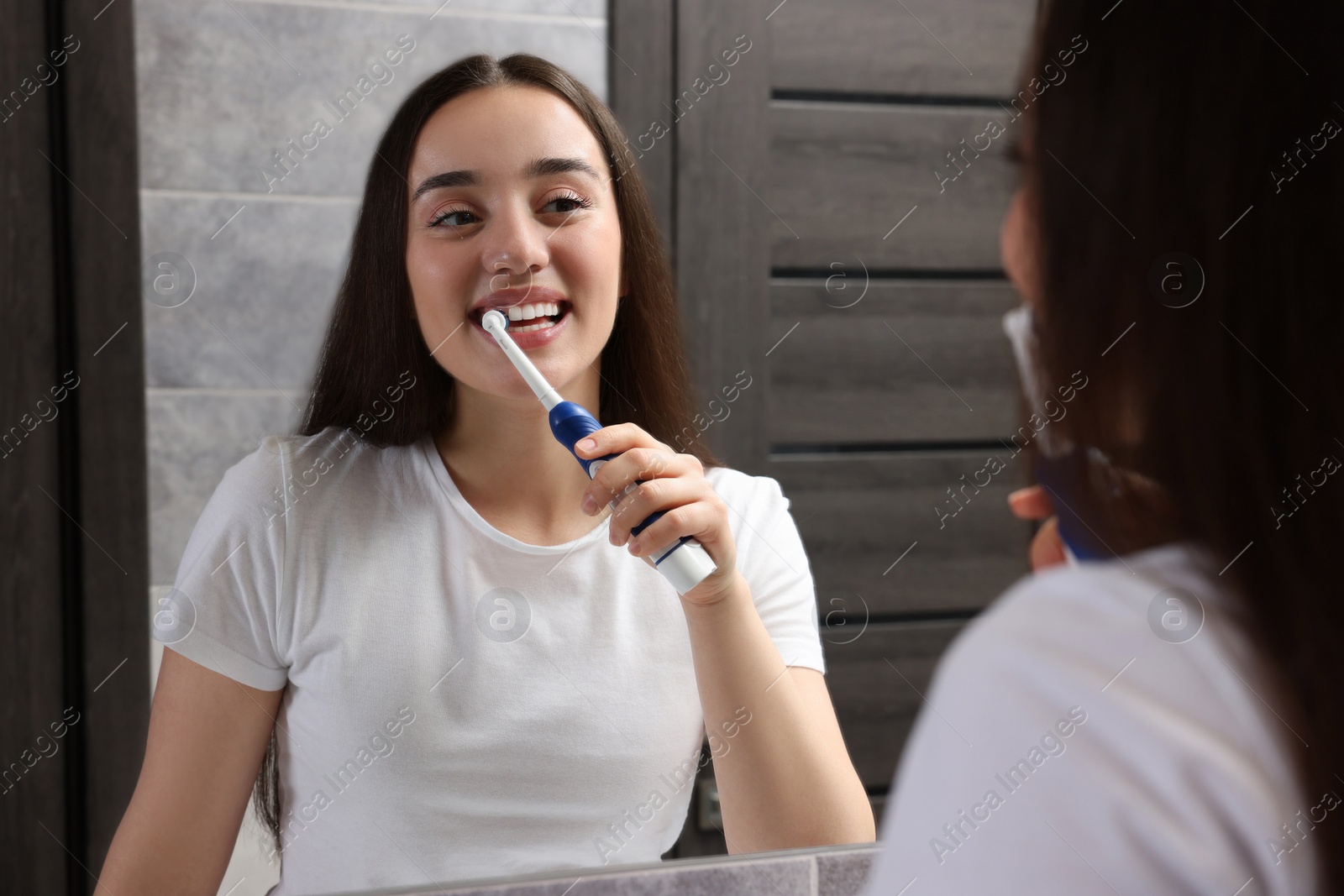 Photo of Young woman brushing her teeth with electric toothbrush near mirror in bathroom