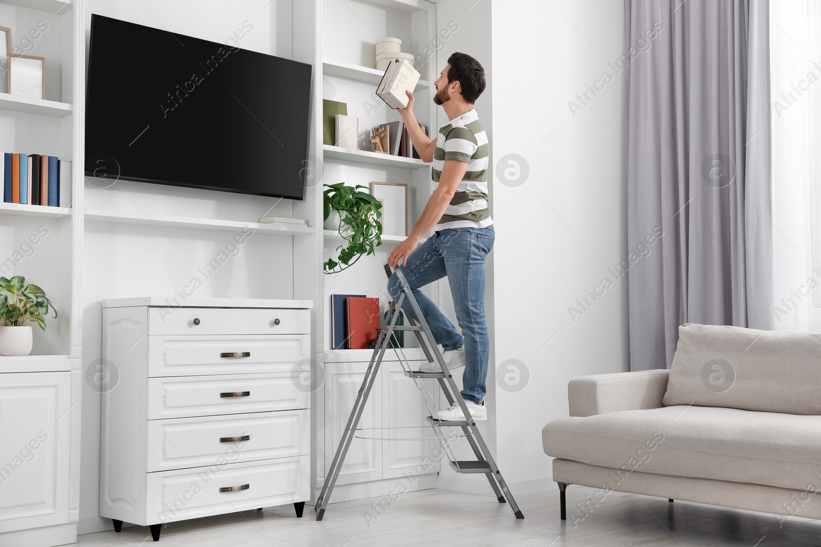 Photo of Man on metal folding ladder taking book from shelf at home