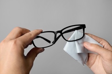 Man wiping glasses with microfiber cloth on light grey background, closeup