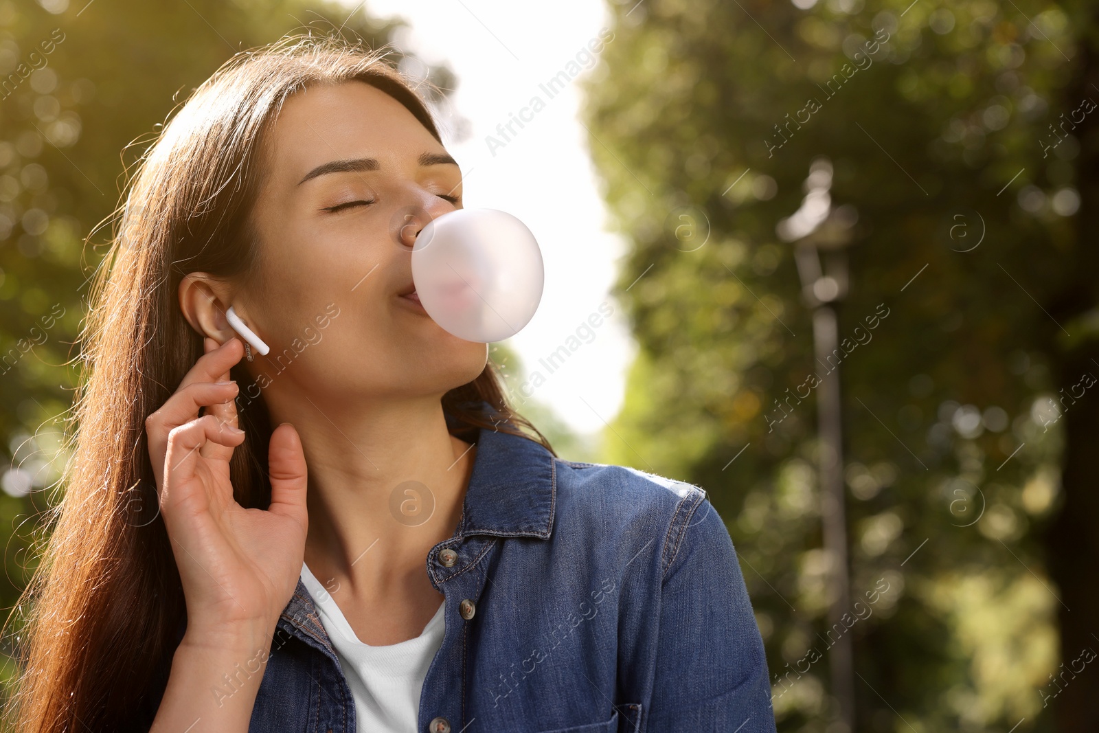 Photo of Beautiful young woman with wireless headphones blowing chewing gum outdoors, space for text