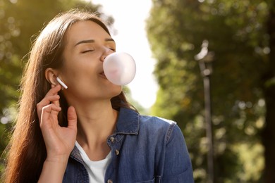 Beautiful young woman with wireless headphones blowing chewing gum outdoors, space for text