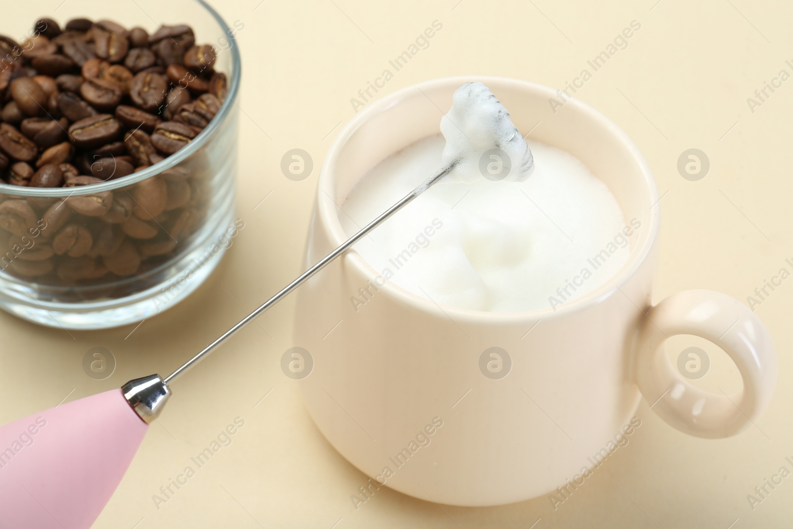 Photo of Mini mixer (milk frother), cup of whipped milk and coffee beans on beige background, closeup