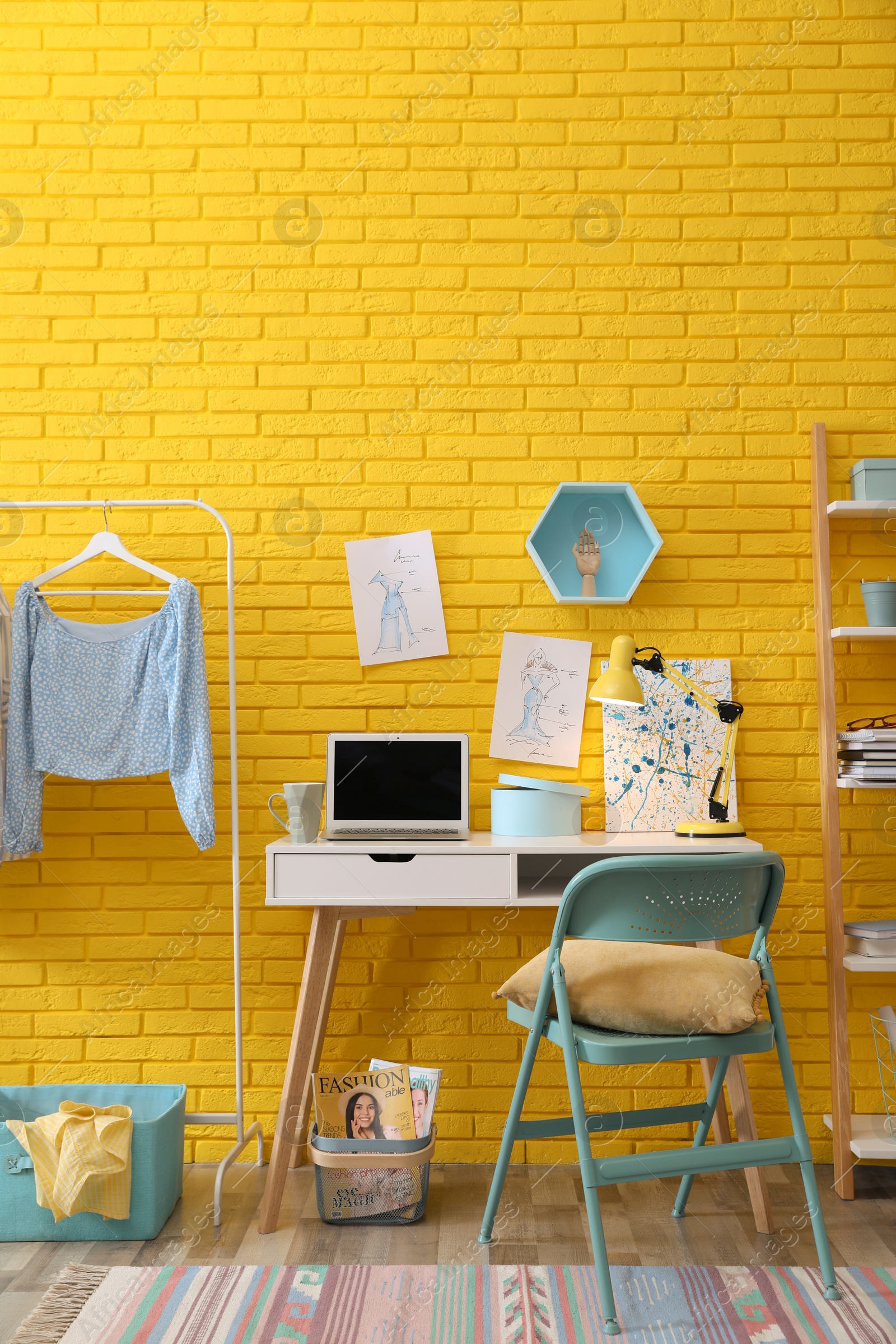 Photo of Fashion designer's workplace with wooden furniture and laptop near yellow brick wall. Stylish interior