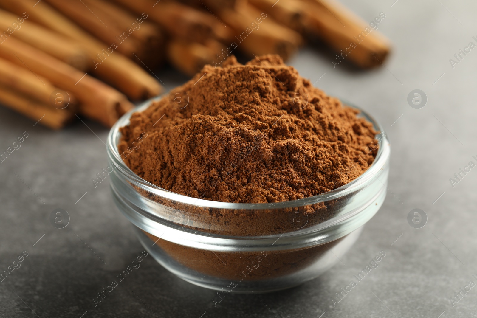 Photo of Bowl of cinnamon powder and sticks on grey table, closeup