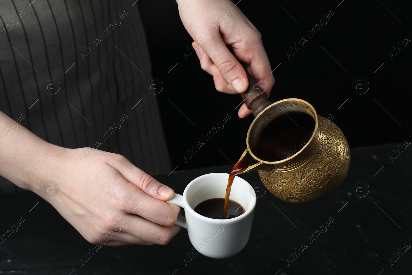 Photo of Turkish coffee. Woman pouring brewed beverage from cezve into cup at black table, closeup