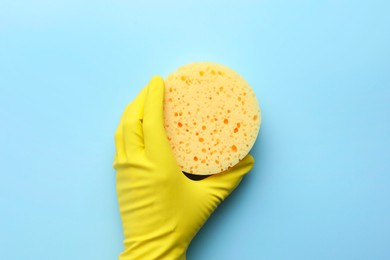 Cleaner in rubber glove holding new yellow sponge on light blue background, top view.