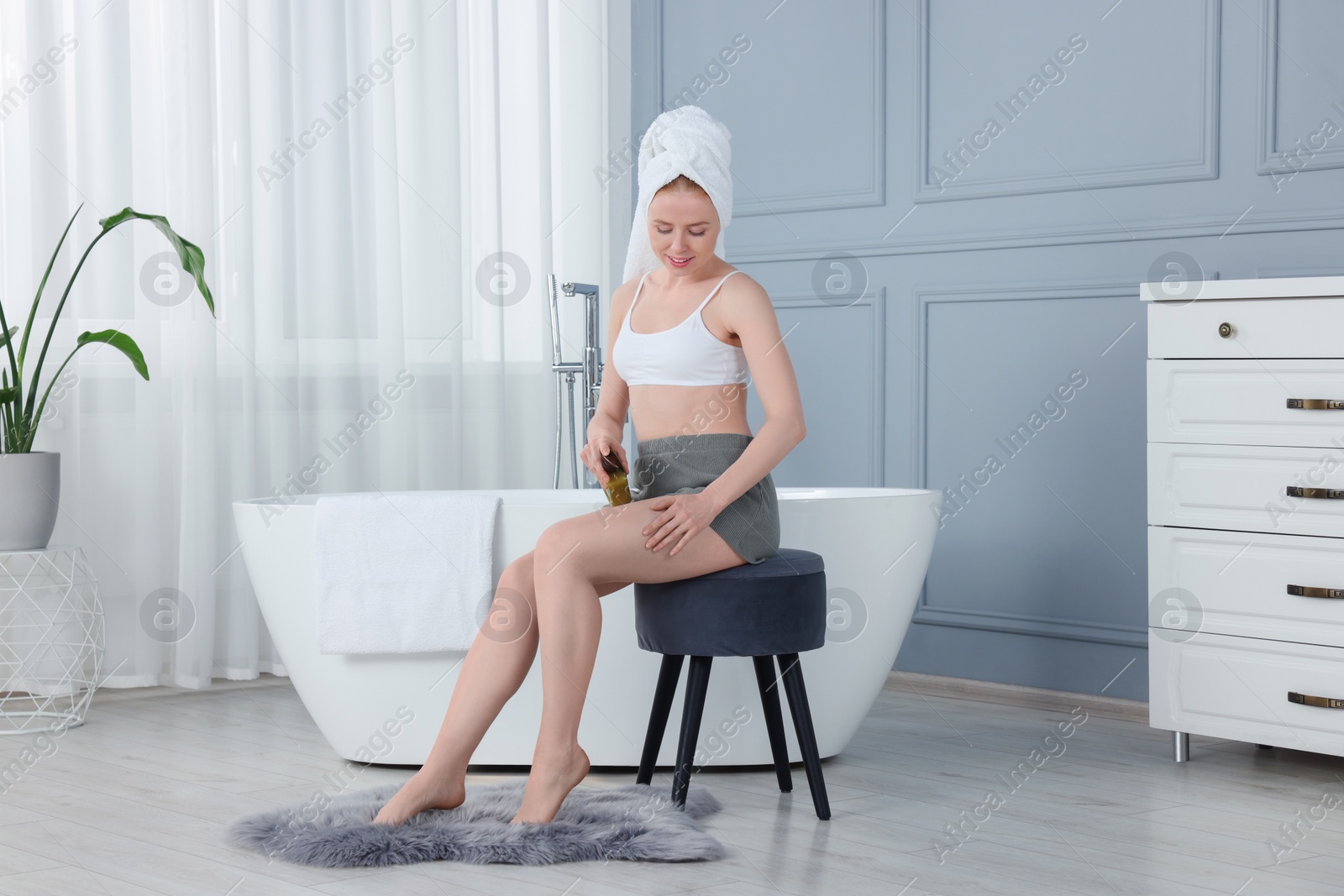Photo of Beautiful young woman applying oil onto leg in bathroom