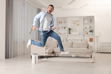 Photo of Enjoying cleaning. Man in headphones jumping with mop at home, space for text
