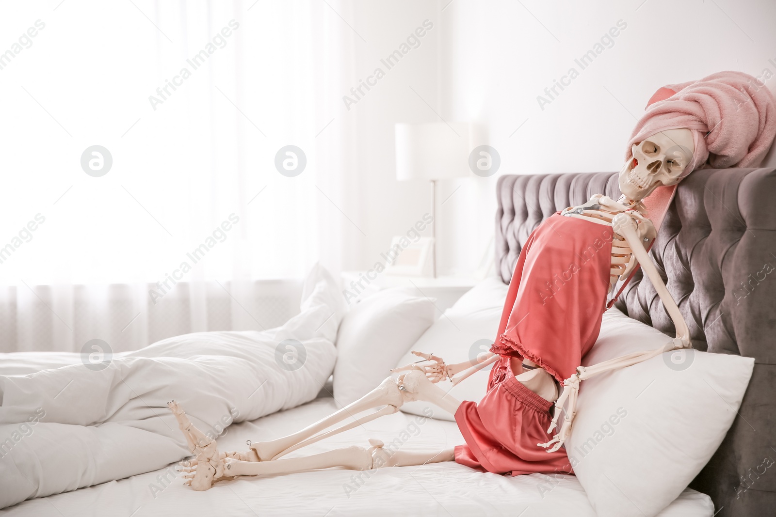 Photo of Human skeleton in silk pajamas and towel sitting on bed indoors