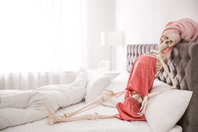 Photo of Human skeleton in silk pajamas and towel sitting on bed indoors