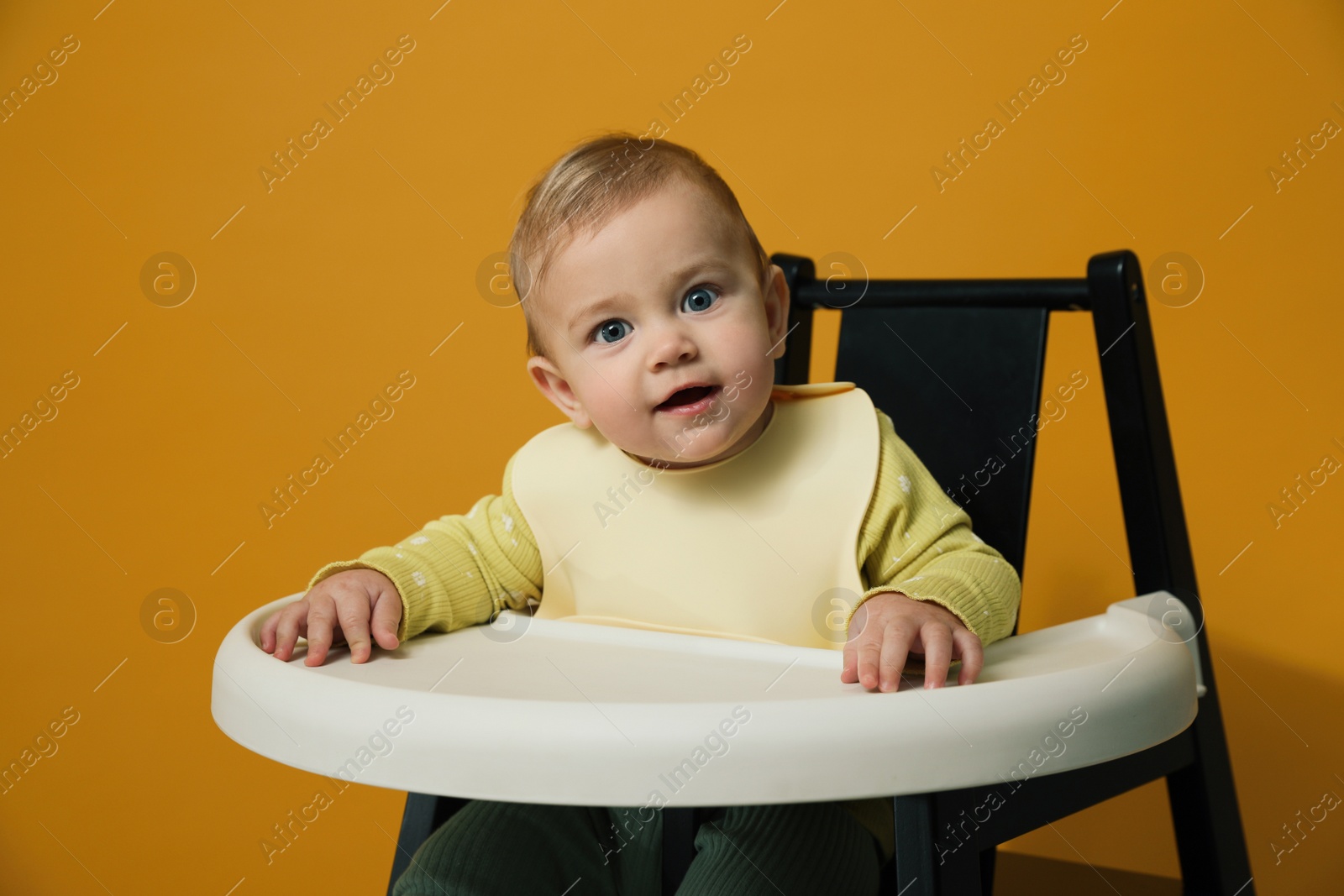 Photo of Cute little baby wearing bib in highchair on yellow background