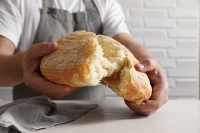 Photo of Man breaking loaf of fresh bread at white table near brick wall, closeup