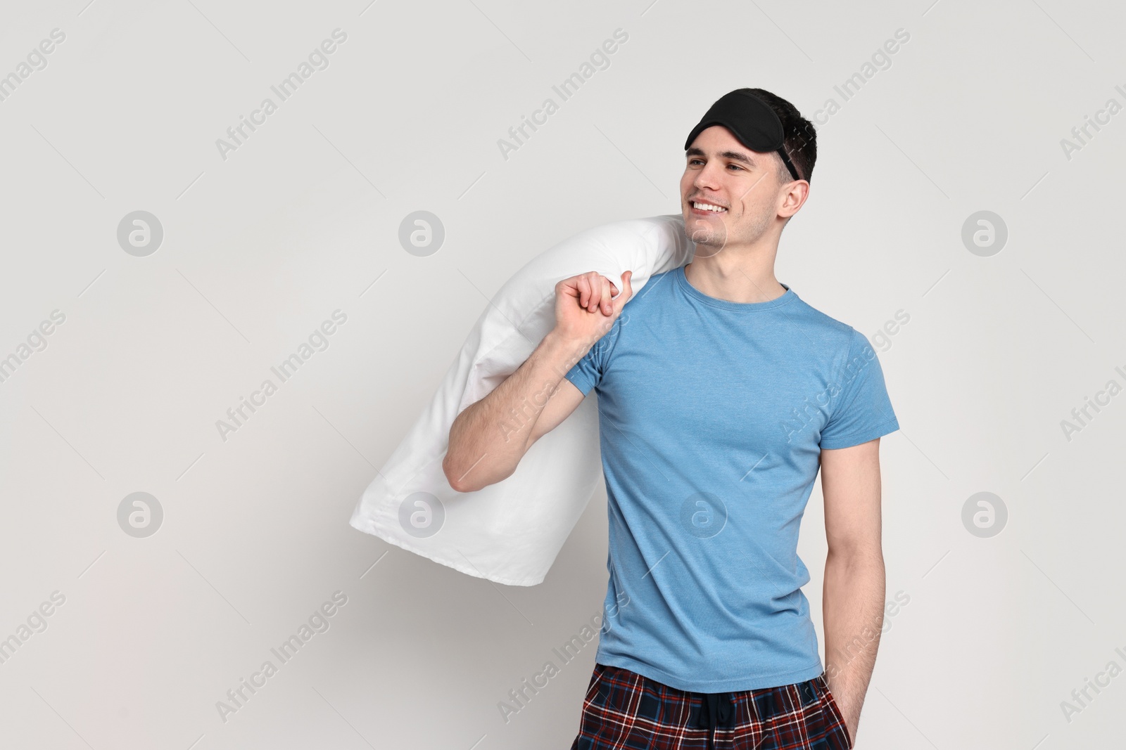 Photo of Happy man in pyjama and sleep mask holding pillow on light grey background, space for text