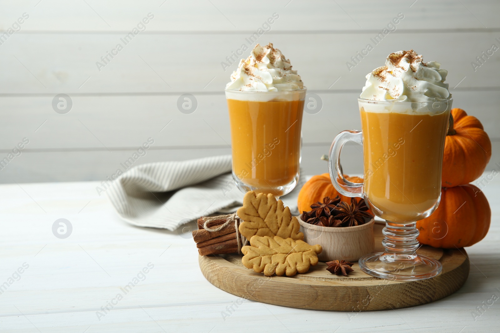 Photo of Tasty pumpkin latte with whipped cream in glasses, spices and cookies on white wooden table. Space for text