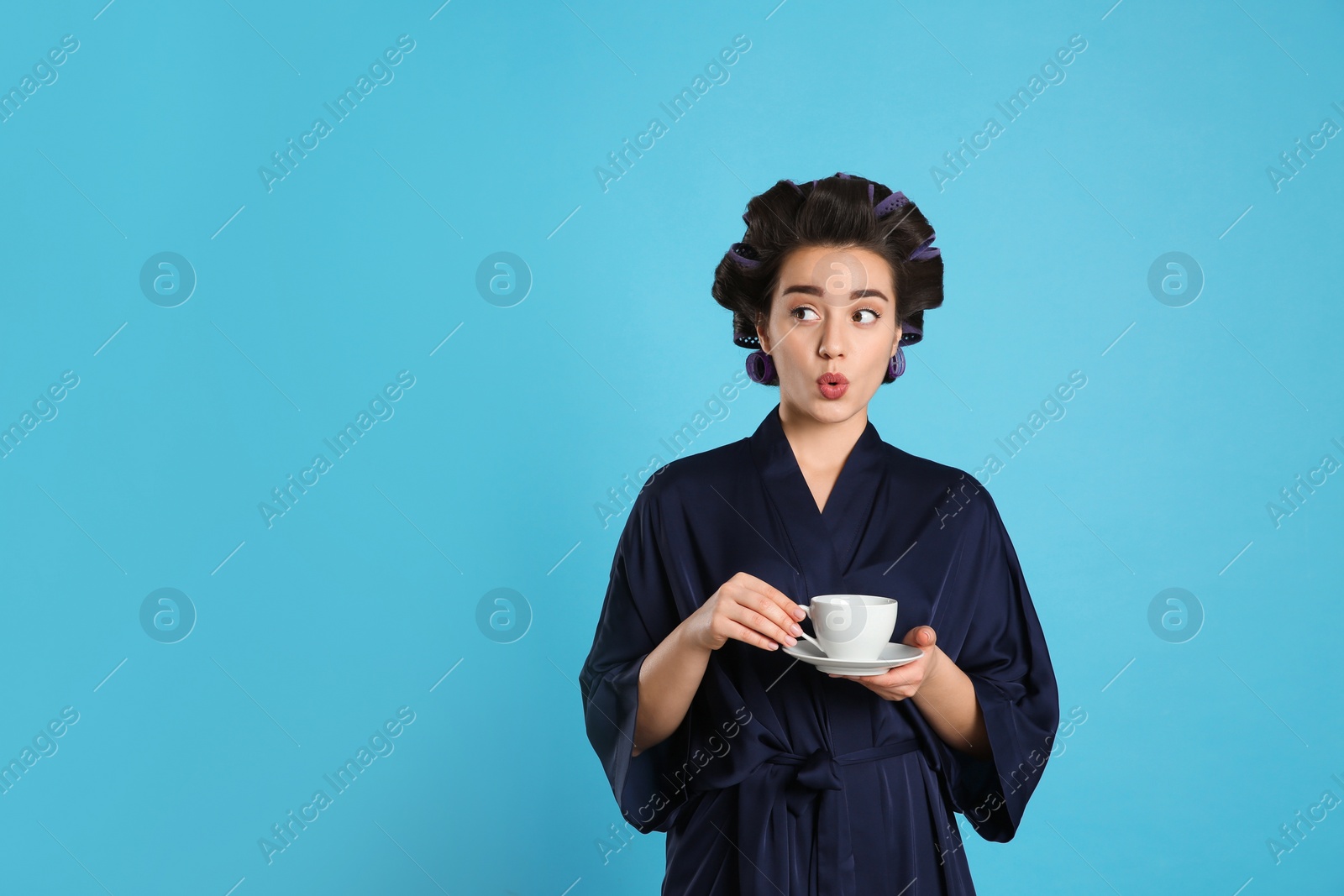 Photo of Emotional young woman in silk bathrobe with hair curlers holding cup of drink on light blue background, space for text