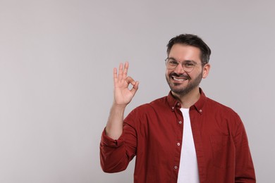 Photo of Portrait of happy man in stylish glasses showing OK gesture on light grey background, space for text
