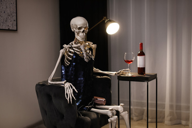 Photo of Skeleton in dress with wine sitting at table indoors
