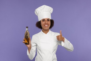 Photo of Happy female chef in uniform holding bottle of cooking oil and showing thumb up on purple background