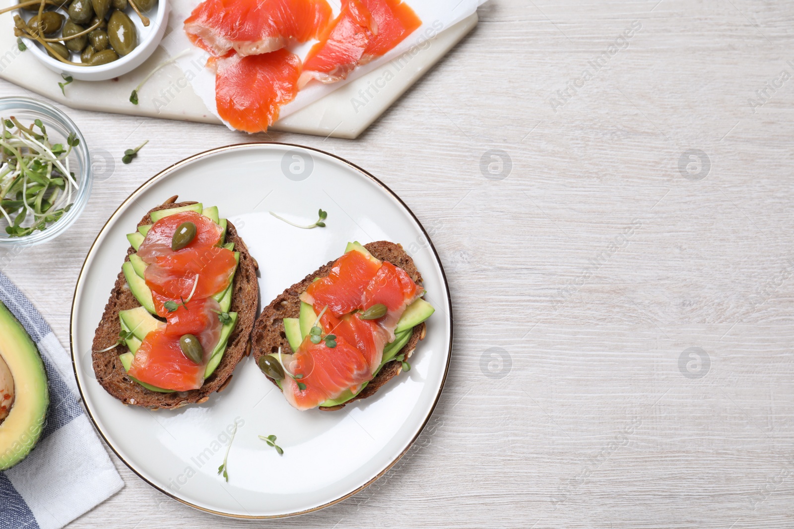 Photo of Delicious sandwiches with salmon, avocado and capers on white wooden table, flat lay. Space for text