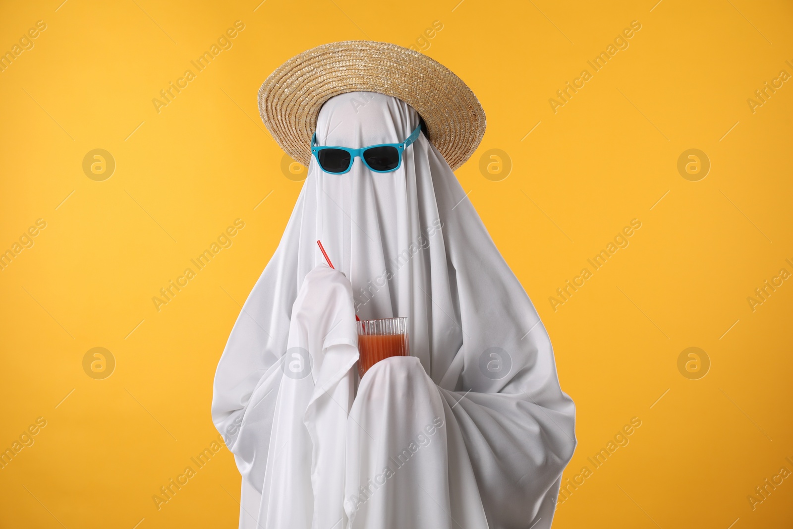 Photo of Person in ghost costume, sunglasses and straw hat holding glass of drink on yellow background