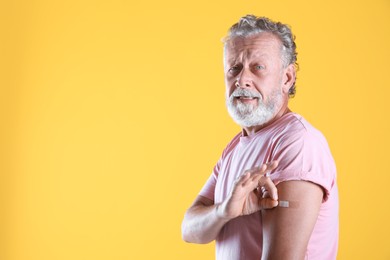 Cheerful senior man showing arm with bandage after vaccination on yellow background. Space for text