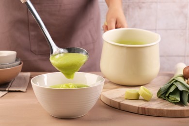 Photo of Woman with ladle pouring tasty leek soup into bowl at wooden table, closeup