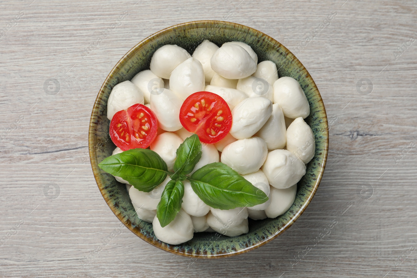 Photo of Delicious mozzarella balls, tomatoes and basil leaves in bowl on white wooden table, top view