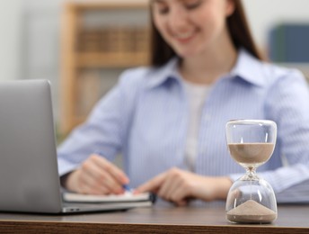 Photo of Hourglass with flowing sand on desk. Woman taking notes while using laptop indoors, selective focus