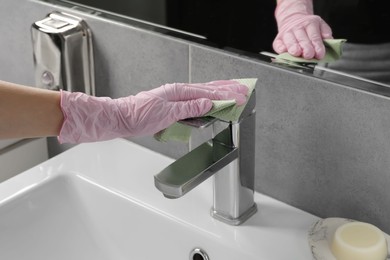 Photo of Woman in glove cleaning faucet of bathroom sink with paper towel, closeup
