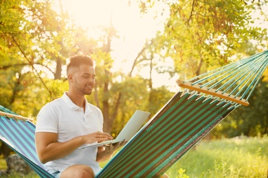 Young man with laptop in comfortable hammock at green garden