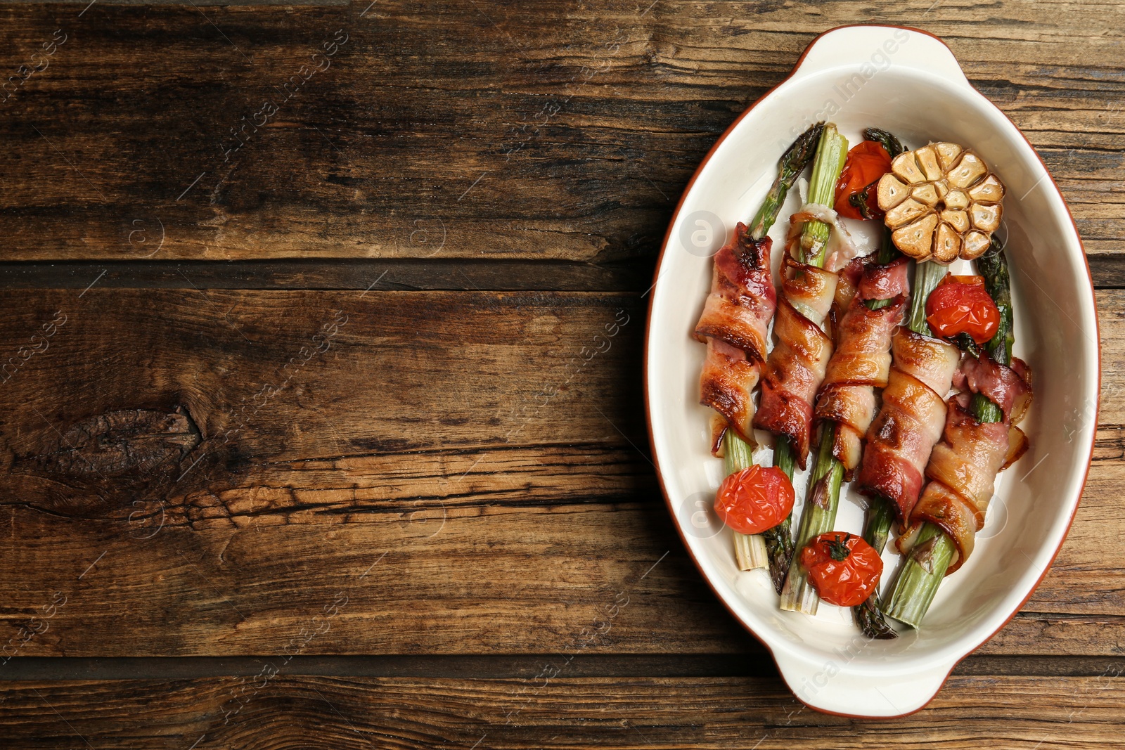 Photo of Oven baked asparagus wrapped with bacon in ceramic dish on wooden table, top view. Space for text