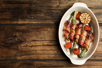 Photo of Oven baked asparagus wrapped with bacon in ceramic dish on wooden table, top view. Space for text