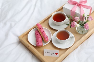 Photo of Tasty breakfast served in bed. Delicious eclairs, tea, gift box, flowers and card with phrase I Love You on tray