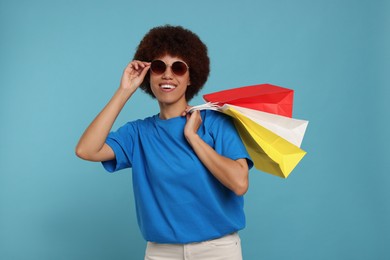 Happy young woman in stylish sunglasses with shopping bags on light blue background